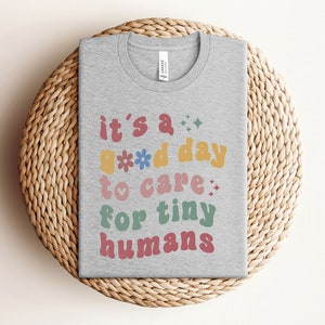 May include: A gray t-shirt with the text "it's a good day to care for tiny humans" in colorful, retro-style lettering.