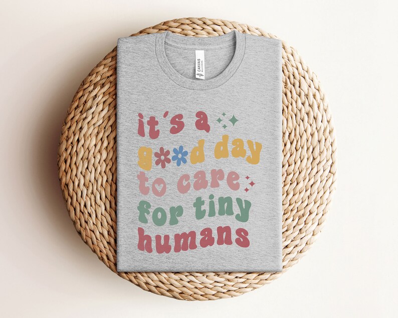 May include: A gray t-shirt with the text "it's a good day to care for tiny humans" in colorful, retro-style lettering.