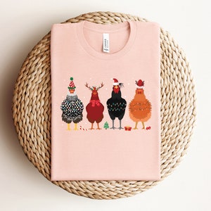 Może przedstawiać: R&oacute;żowy t-shirt z grafiką przedstawiającą cztery kury w świątecznych czapkach i dodatkach. Kury są ustawione w rzędzie i patrzą w stronę widza. T-shirt leży na plecionym słomianym dywaniku.