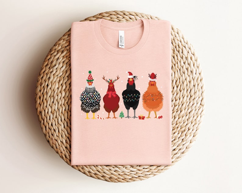 Może przedstawiać: R&oacute;żowy t-shirt z grafiką przedstawiającą cztery kury w świątecznych czapkach i dodatkach. Kury są ustawione w rzędzie i patrzą w stronę widza. T-shirt leży na plecionym słomianym dywaniku.