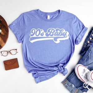 Op de afbeelding: Een heather columbia blauw t-shirt met de tekst "90's Baby" in een wit retro lettertype.