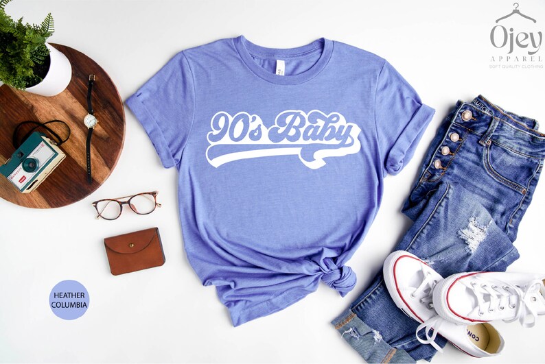 Op de afbeelding: Een heather columbia blauw t-shirt met de tekst "90's Baby" in een wit retro lettertype.