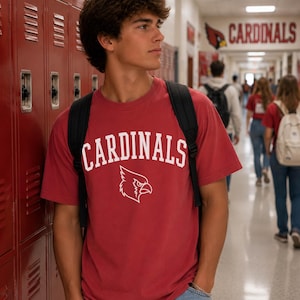 May include: A red short-sleeved t-shirt featuring the word CARDINALS in white arched lettering above a white graphic of a cardinal bird head. The shirt is worn with a black backpack and light blue denim jeans, creating a casual look.