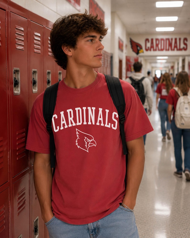 May include: A red short-sleeved t-shirt featuring the word CARDINALS in white arched lettering above a white graphic of a cardinal bird head. The shirt is worn with a black backpack and light blue denim jeans, creating a casual look.