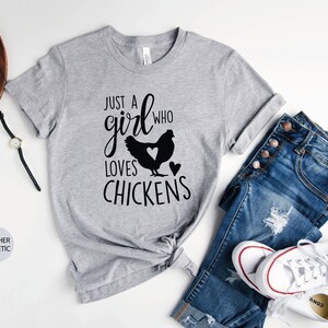 Puede incluir: Una camiseta gris jaspeada con el texto "Just a girl who loves chickens" en letras negras. La camiseta presenta una silueta de pollo con un coraz&oacute;n en el pecho.