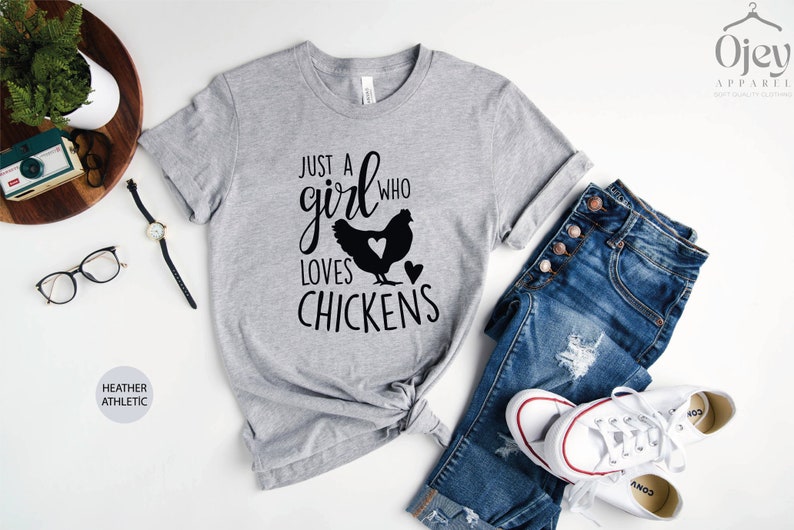 Puede incluir: Una camiseta gris jaspeada con el texto "Just a girl who loves chickens" en letras negras. La camiseta presenta una silueta de pollo con un coraz&oacute;n en el pecho.