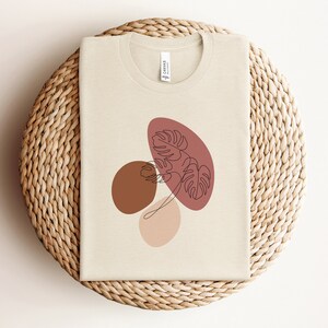Puede incluir: Una camiseta beige con un dise&ntilde;o gr&aacute;fico de formas abstractas en tonos marrones, rosas y rojos. El dise&ntilde;o presenta una hoja estilizada en el centro.