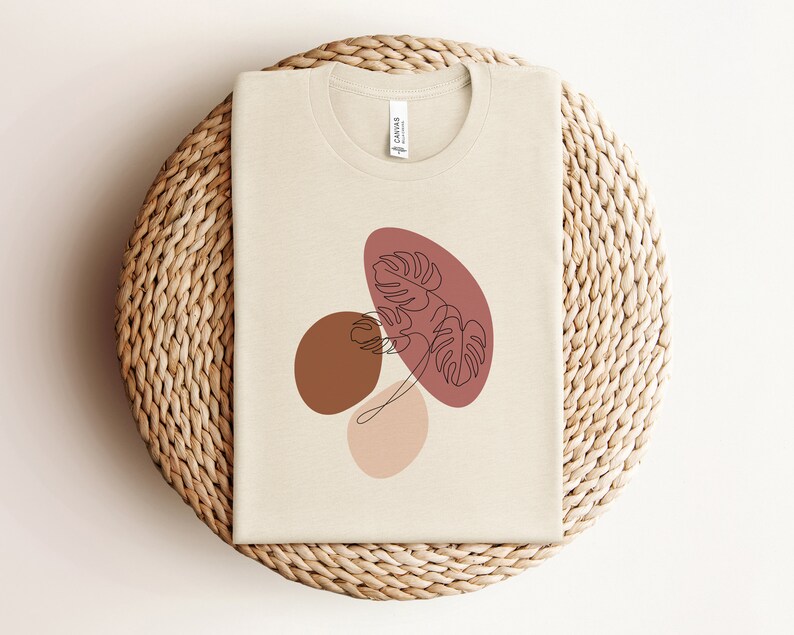 Puede incluir: Una camiseta beige con un dise&ntilde;o gr&aacute;fico de formas abstractas en tonos marrones, rosas y rojos. El dise&ntilde;o presenta una hoja estilizada en el centro.