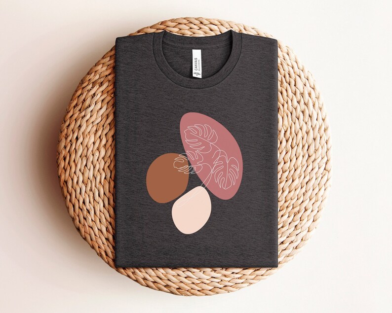 Puede incluir: Una camiseta gris oscuro con un dise&ntilde;o gr&aacute;fico de formas abstractas en tonos de rosa, marr&oacute;n y blanco. El dise&ntilde;o presenta una hoja estilizada con un dibujo lineal de una planta.