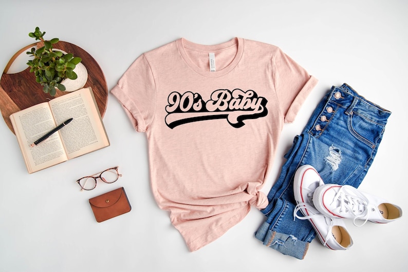 Op de afbeelding: Een roze T-shirt met de tekst "90's Baby" in een retro zwart lettertype. Het shirt is gestyled met een blauwe jeans en witte sneakers.