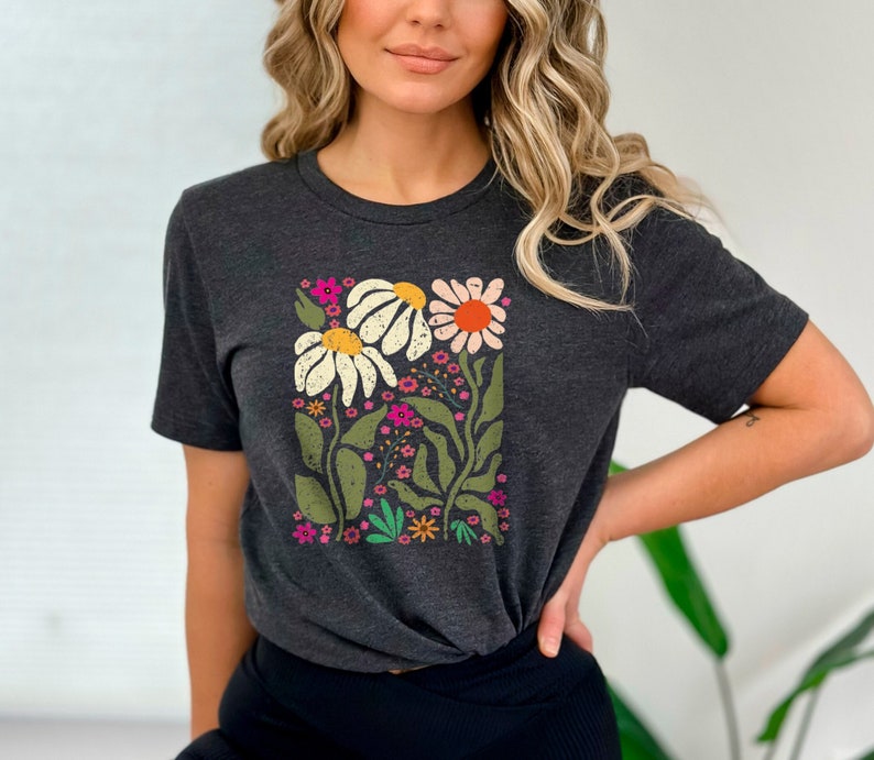 May include: A dark gray t-shirt with a floral print featuring white, pink, orange, and green flowers. The flowers are arranged in a square shape on the front of the shirt.