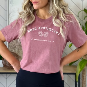 May include: A dusty rose-colored t-shirt with the text "ROSE APOTHECARY" in white, arched above two floral illustrations. Below the flowers, the text "HANDCRAFTED WITH CARE" is printed. The shirt is a crew neck style.