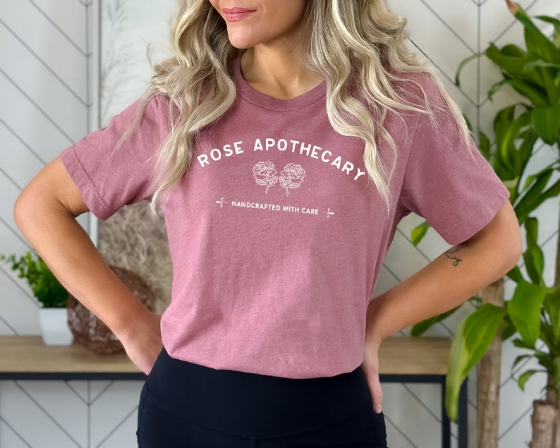 May include: A dusty rose-colored t-shirt with the text "ROSE APOTHECARY" in white, arched above two floral illustrations. Below the flowers, the text "HANDCRAFTED WITH CARE" is printed. The shirt is a crew neck style.