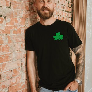 May include: A man wearing a black t-shirt with a green shamrock graphic. He is standing in front of a brick wall and has a beard.