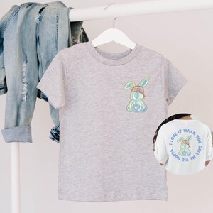 Puede incluir: Una camiseta gris con un conejito de dibujos animados colorido que lleva un sombrero y gafas de sol. La parte posterior de la camiseta tiene el texto "Me encanta cuando me llamas mami grande".