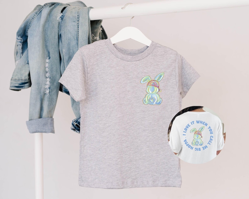 Puede incluir: Una camiseta gris con un conejito de dibujos animados colorido que lleva un sombrero y gafas de sol. La parte posterior de la camiseta tiene el texto "Me encanta cuando me llamas mami grande".