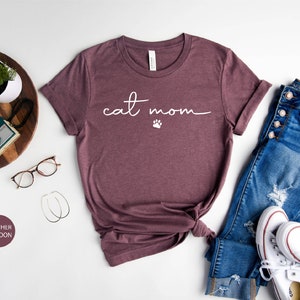 May include: Heather maroon t-shirt with the text "cat mom" in white script with a paw print. The shirt is laid out on a white surface with a pair of glasses, a wooden tray with a camera and watch, a pair of hoop earrings, and a pair of blue jeans with white sneakers.