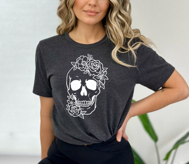 Peut inclure: Un t-shirt gris fonc&eacute; avec un motif de cr&acirc;ne floral blanc. Le cr&acirc;ne est entour&eacute; de fleurs et de feuilles.