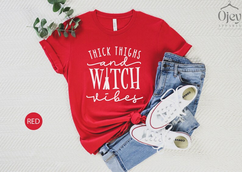 May include: Red t-shirt with the text "Thick Thighs and Witch Vibes" in white lettering. The shirt is laid out on a white surface with a pair of blue jeans and white sneakers.