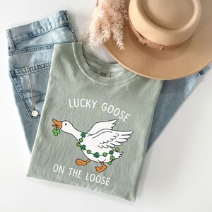 May include: Sage green t-shirt with a graphic of a white goose wearing a shamrock garland and holding a shamrock in its beak. The text "LUCKY GOOSE ON THE LOOSE" is printed above and below the goose. Light blue jeans and a tan hat are in the background.