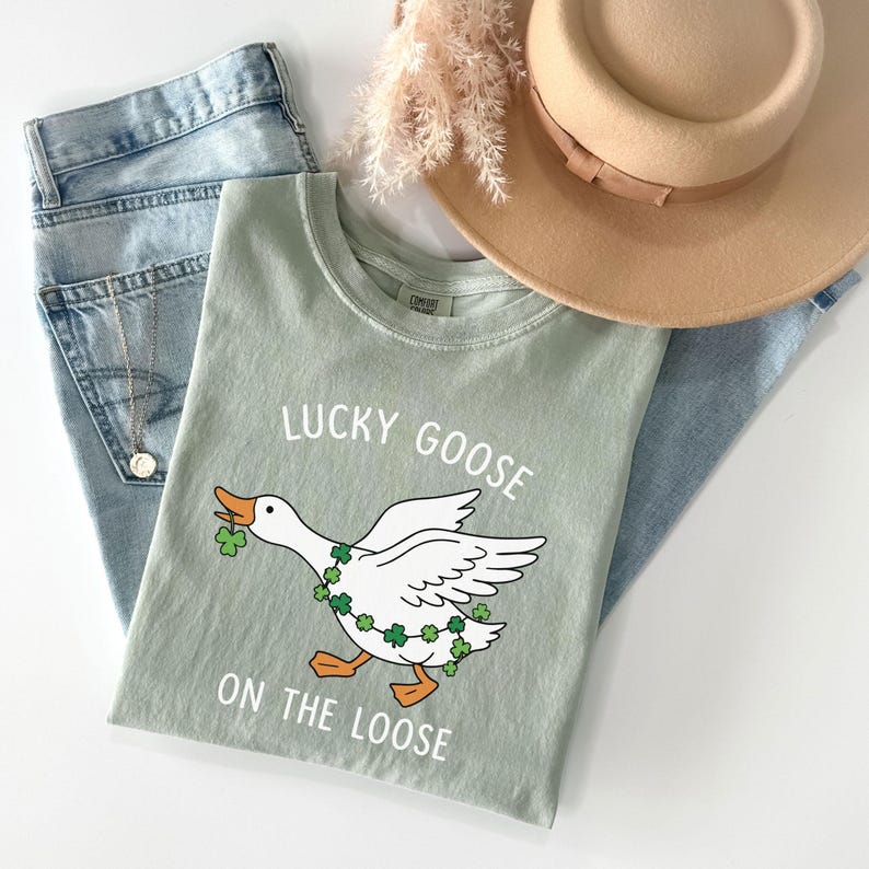 May include: Sage green t-shirt with a graphic of a white goose wearing a shamrock garland and holding a shamrock in its beak. The text "LUCKY GOOSE ON THE LOOSE" is printed above and below the goose. Light blue jeans and a tan hat are in the background.