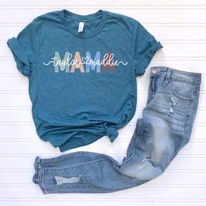 May include: Teal-colored t-shirt with the word "MAMA" in pastel colors and the names "Taylor" and "Maddie" in a cursive font. Light blue denim jeans with distressed details are next to the shirt. The items are arranged on a white surface.