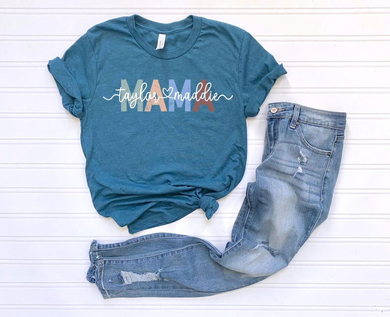 May include: Teal-colored t-shirt with the word "MAMA" in pastel colors and the names "Taylor" and "Maddie" in a cursive font. Light blue denim jeans with distressed details are next to the shirt. The items are arranged on a white surface.