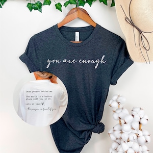 May include: A dark grey t-shirt with the text "you are enough" printed in white on the front. The back of the shirt has a white message that reads "Dear person behind me: The world is a better place with you in it. Lots of love, -the person in front of you".