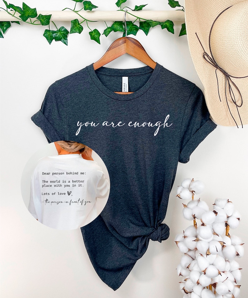 May include: A dark grey t-shirt with the text "you are enough" printed in white on the front. The back of the shirt has a white message that reads "Dear person behind me: The world is a better place with you in it. Lots of love, -the person in front of you".