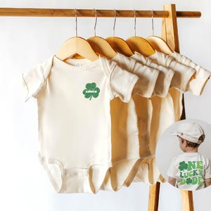K&ouml;nnte beinhalten: F&uuml;nf cremefarbene Baby-Bodys mit kurzen &Auml;rmeln h&auml;ngen an Holzb&uuml;geln. Der Body links hat ein gr&uuml;nes Kleeblatt mit dem Namen "Andrew" darauf gedruckt. Im Hintergrund ist ein Kind zu sehen, das ein wei&szlig;es T-Shirt mit einem gr&uuml;nen Kleeblatt und dem Text "One Lucky Dude" tr&auml;gt.