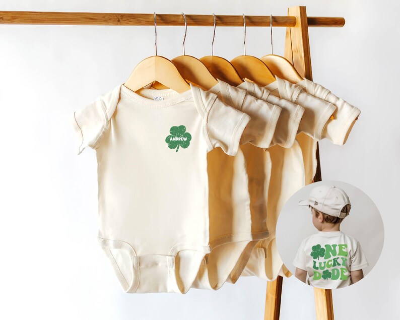 K&ouml;nnte beinhalten: F&uuml;nf cremefarbene Baby-Bodys mit kurzen &Auml;rmeln h&auml;ngen an Holzb&uuml;geln. Der Body links hat ein gr&uuml;nes Kleeblatt mit dem Namen "Andrew" darauf gedruckt. Im Hintergrund ist ein Kind zu sehen, das ein wei&szlig;es T-Shirt mit einem gr&uuml;nen Kleeblatt und dem Text "One Lucky Dude" tr&auml;gt.