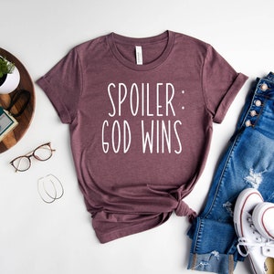 May include: A mauve t-shirt with the text "Spoiler: God Wins" in white lettering. The shirt is laid out on a white surface with a pair of jeans, a pair of white trainers, a potted plant, a camera, and a pair of glasses.