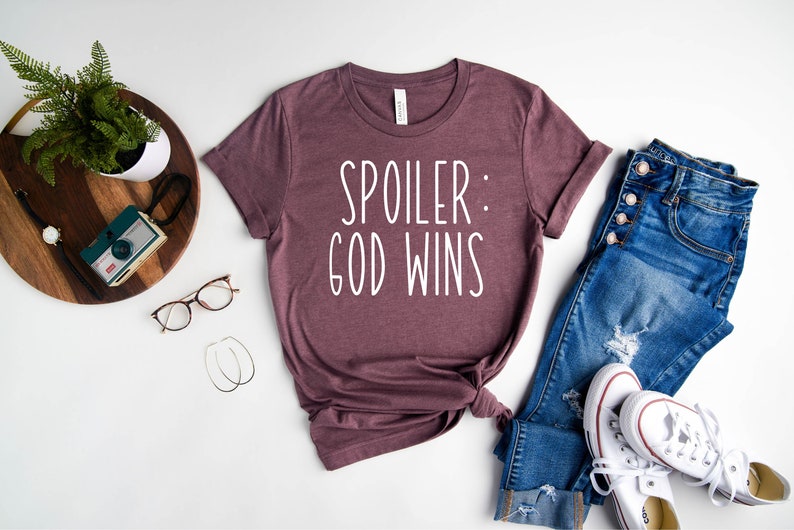 May include: A mauve t-shirt with the text "Spoiler: God Wins" in white lettering. The shirt is laid out on a white surface with a pair of jeans, a pair of white trainers, a potted plant, a camera, and a pair of glasses.