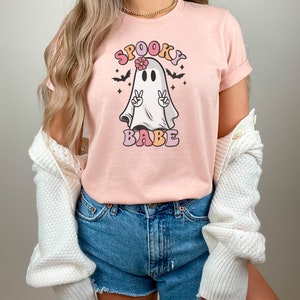 May include: A pink t-shirt with a cartoon ghost wearing a flower and making a peace sign. The text "Spooky Babe" is printed in colorful letters below the ghost.