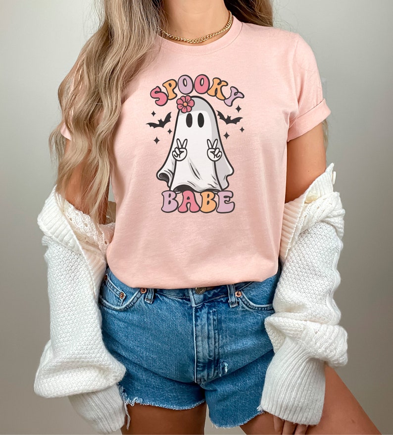 May include: A pink t-shirt with a cartoon ghost wearing a flower and making a peace sign. The text "Spooky Babe" is printed in colorful letters below the ghost.