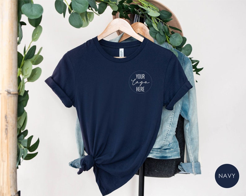 May include: Navy blue t-shirt with rolled sleeves, hanging on a wooden hanger. The shirt features a circular logo design with the text "Your logo here". A denim jacket hangs behind the shirt. The word "NAVY" is in a navy blue circle.