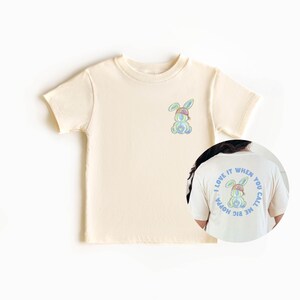 Puede incluir: Una camiseta de color crema con un conejito de dibujos animados con un sombrero en la parte delantera. La parte posterior de la camiseta tiene un c&iacute;rculo con el texto "Me encanta cuando me llamas gran hoppa" y un conejito de dibujos animados con un sombrero dentro del c&iacute;rculo.