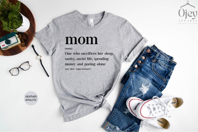 Może przedstawiać: Szary t-shirt z nadrukiem słowa "mom" zdefiniowanego jako "Osoba, kt&oacute;ra poświęca sw&oacute;j sen, zdrowie psychiczne, życie towarzyskie, pieniądze i oddaje mocz sama (patrz także: 'superwoman')".
