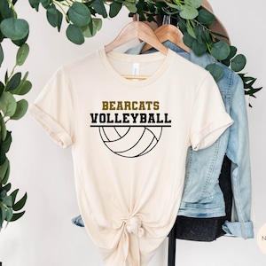 K&ouml;nnte beinhalten: Ein beiges T-Shirt mit dem Text "Bearcats Volleyball" in schwarzer und goldener Schrift, darunter eine Grafik eines schwarzen und wei&szlig;en Volleyballs.