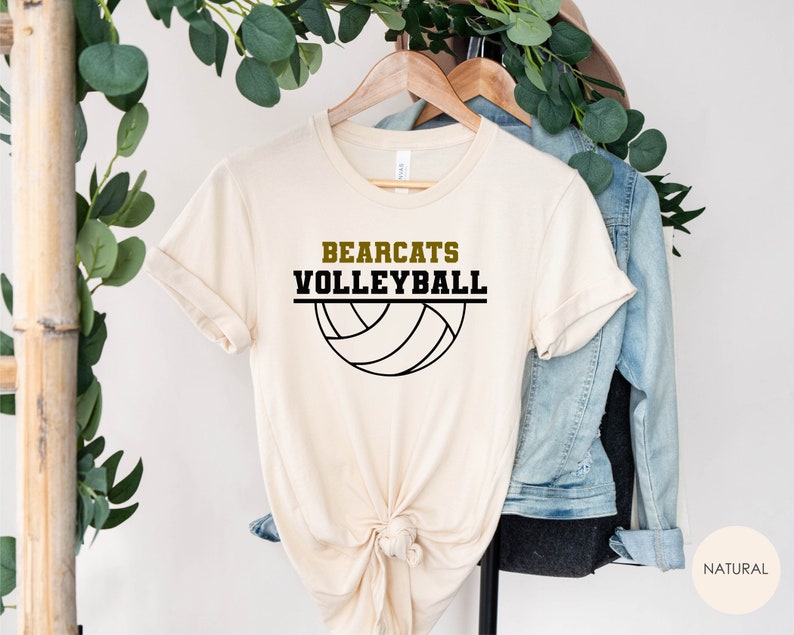 K&ouml;nnte beinhalten: Ein beiges T-Shirt mit dem Text "Bearcats Volleyball" in schwarzer und goldener Schrift, darunter eine Grafik eines schwarzen und wei&szlig;en Volleyballs.