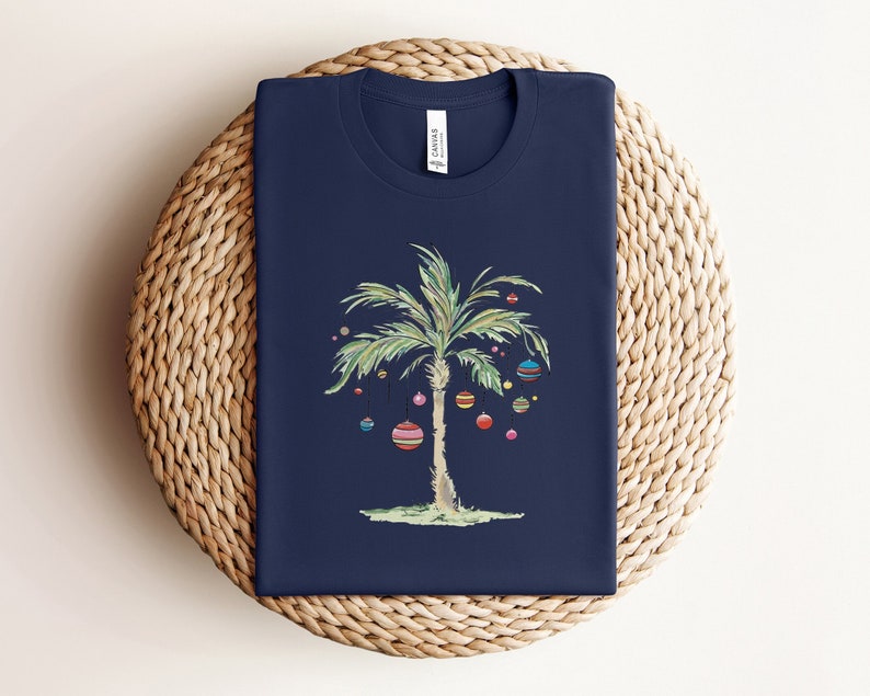 K&ouml;nnte beinhalten: Ein marineblaues T-Shirt mit einem wei&szlig;en Palmenbaum-Design mit bunten Ornamenten, die an den &Auml;sten h&auml;ngen. Der Palmenbaum ist mittig auf dem T-Shirt platziert.