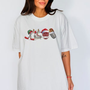 May include: White oversized t-shirt with a Christmas-themed hockey design. The design features hockey sticks, skates, a helmet, and a puck, all decorated with Christmas lights and Santa hats.