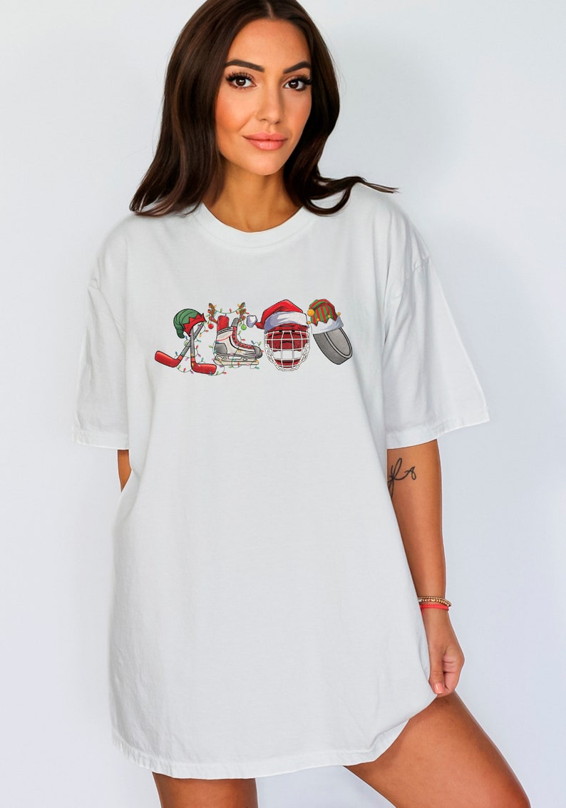 May include: White oversized t-shirt with a Christmas-themed hockey design. The design features hockey sticks, skates, a helmet, and a puck, all decorated with Christmas lights and Santa hats.