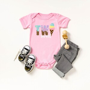 May include: Pink baby onesie with the text "Two Sweet" and an ice cream cone graphic. The onesie is paired with gray jeans, black sneakers, and a wooden rattle.