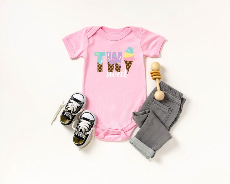 May include: Pink baby onesie with the text "Two Sweet" and an ice cream cone graphic. The onesie is paired with gray jeans, black sneakers, and a wooden rattle.