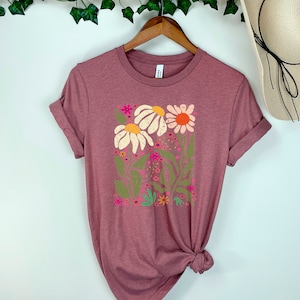 May include: A mauve t-shirt with a floral print featuring white, pink, orange, and green flowers. The shirt has a crew neck and short sleeves.