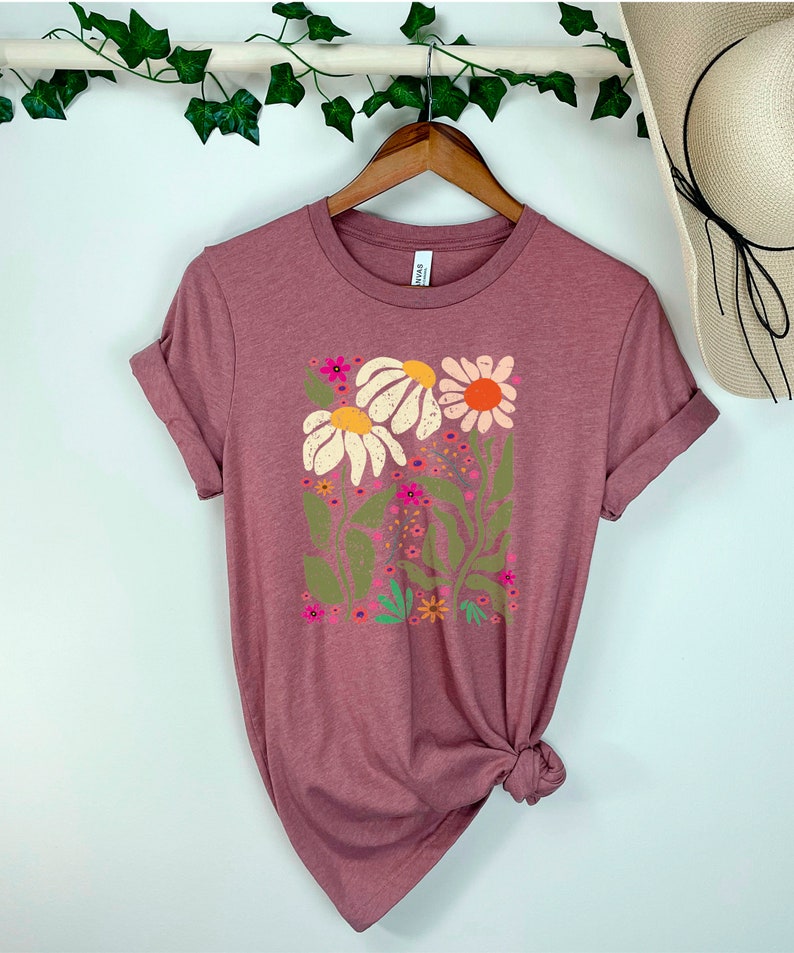 May include: A mauve t-shirt with a floral print featuring white, pink, orange, and green flowers. The shirt has a crew neck and short sleeves.