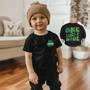 K&ouml;nnte beinhalten: Ein kleines Kind tr&auml;gt ein schwarzes T-Shirt mit einem gr&uuml;nen Kleeblatt und dem Namen "Jackson" darauf. Das Kind tr&auml;gt auch eine braune M&uuml;tze und eine schwarze Hose. Das T-Shirt hat ein gr&uuml;nes Kleeblatt-Design mit den Worten "One Lucky Dude" darauf.