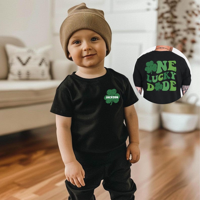 K&ouml;nnte beinhalten: Ein kleines Kind tr&auml;gt ein schwarzes T-Shirt mit einem gr&uuml;nen Kleeblatt und dem Namen "Jackson" darauf. Das Kind tr&auml;gt auch eine braune M&uuml;tze und eine schwarze Hose. Das T-Shirt hat ein gr&uuml;nes Kleeblatt-Design mit den Worten "One Lucky Dude" darauf.