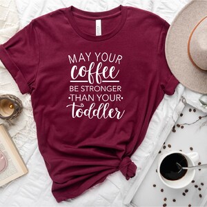 May include: A maroon t-shirt with the text "May your coffee be stronger than your toddler" in white lettering.